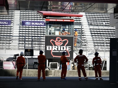A group of people in orange uniforms and helmets are standing in front of a grandstand. In the background, there is a large advertising billboard with the word 'BRIDE' prominently displayed. The stadium seats are mostly empty, and several individuals are visible on a staircase near the sign.