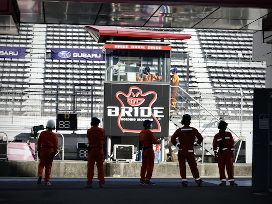 A group of people in orange uniforms and helmets are standing in front of a grandstand. In the background, there is a large advertising billboard with the word 'BRIDE' prominently displayed. The stadium seats are mostly empty, and several individuals are visible on a staircase near the sign.