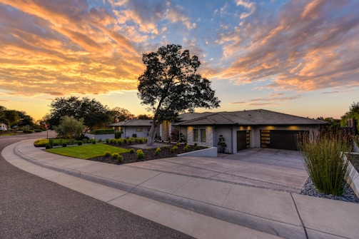 A modern suburban house with a well-manicured lawn and a large tree in the front yard. The driveway leads to a spacious garage, and the sky above is painted with a beautiful sunset, featuring vibrant shades of orange, pink, and blue. The street is quiet and clean, curving just in front of the house.