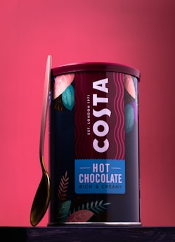 A container of Costa hot chocolate with a rich and creamy tagline is prominently displayed. The packaging is adorned with colorful leaves, and a gold-colored spoon leans against the container. A red background complements the vibrant branding on the can.