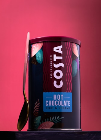 A container of Costa hot chocolate with a rich and creamy tagline is prominently displayed. The packaging is adorned with colorful leaves, and a gold-colored spoon leans against the container. A red background complements the vibrant branding on the can.