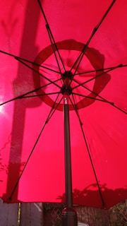 A red umbrella is opened from underneath, showing its black spokes and central pole. Sunlight creates shadows and highlights the texture of the fabric. The background includes hints of a wooden fence and foliage.