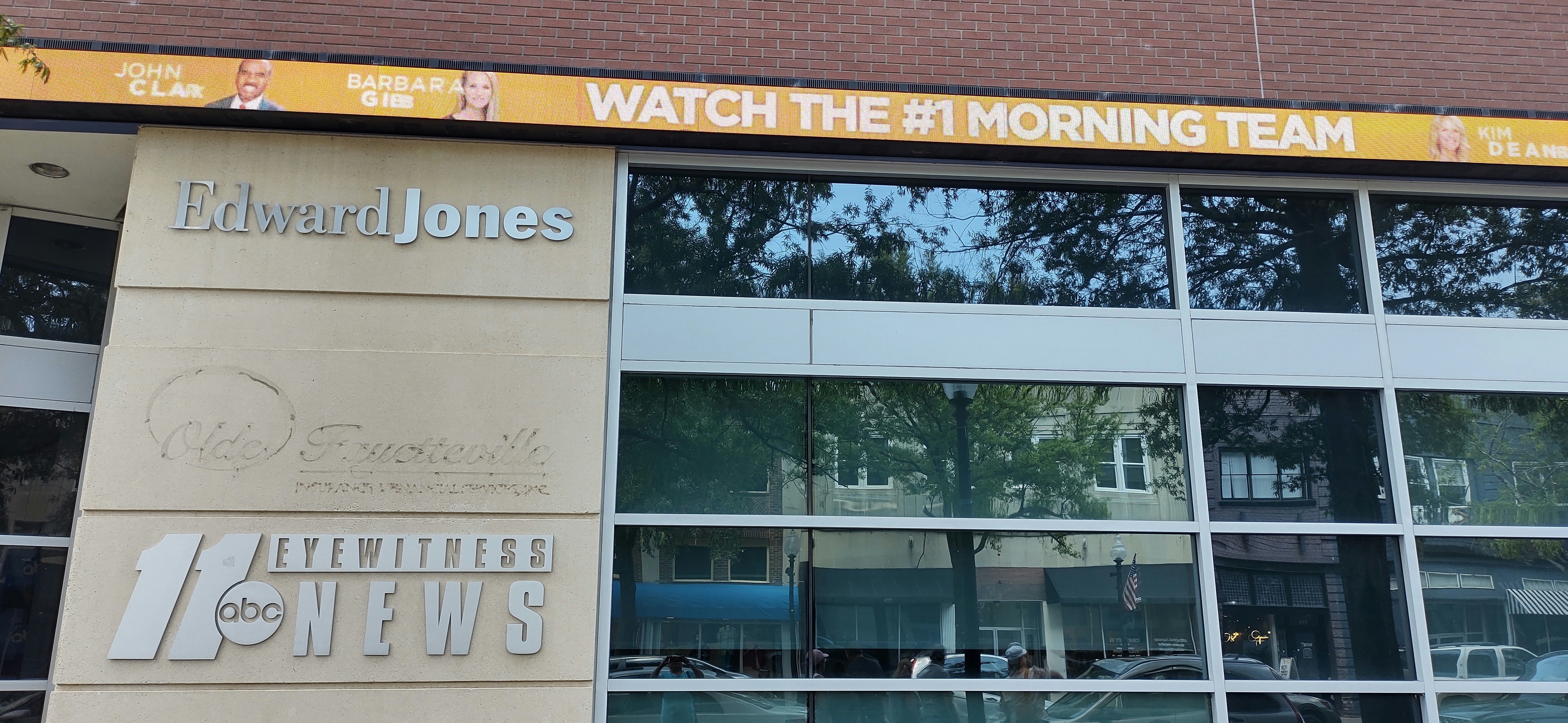 The image shows a building with large windows and multiple signs. One sign reads 'Edward Jones' in silver letters, while another displays the logo and text for '11 abc Eyewitness News.' Above the building, a digital banner promotes a morning news team with images of three individuals and the text 'WATCH THE #1 MORNING TEAM.'