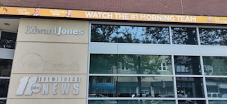 The image shows a building with large windows and multiple signs. One sign reads 'Edward Jones' in silver letters, while another displays the logo and text for '11 abc Eyewitness News.' Above the building, a digital banner promotes a morning news team with images of three individuals and the text 'WATCH THE #1 MORNING TEAM.'