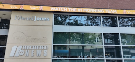 The image shows a building with large windows and multiple signs. One sign reads 'Edward Jones' in silver letters, while another displays the logo and text for '11 abc Eyewitness News.' Above the building, a digital banner promotes a morning news team with images of three individuals and the text 'WATCH THE #1 MORNING TEAM.'