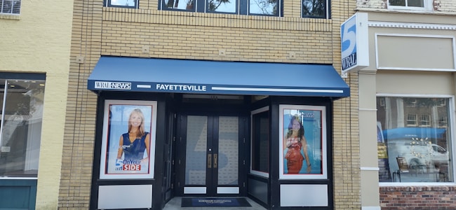 The image features the entrance of a building with a dark double door flanked by two large posters displaying individuals. Above the entrance is a blue awning with 'WRAL NEWS FAYETTEVILLE' written on it. The building is made of light brown bricks and has a modern appearance.