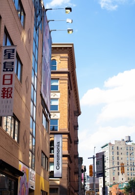 Several multi-story buildings with signs in multiple languages line a city street. The sky is clear with scattered clouds. One prominent sign reads 'Ceremonia', while others feature Asian characters.