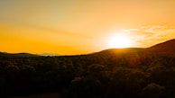 A vibrant sunset over a dense forest with a bright sun dipping behind the hills. The sky is painted in warm hues, with a gradient of orange and yellow. Gentle clouds stretch across the sky, adding texture to the scene.