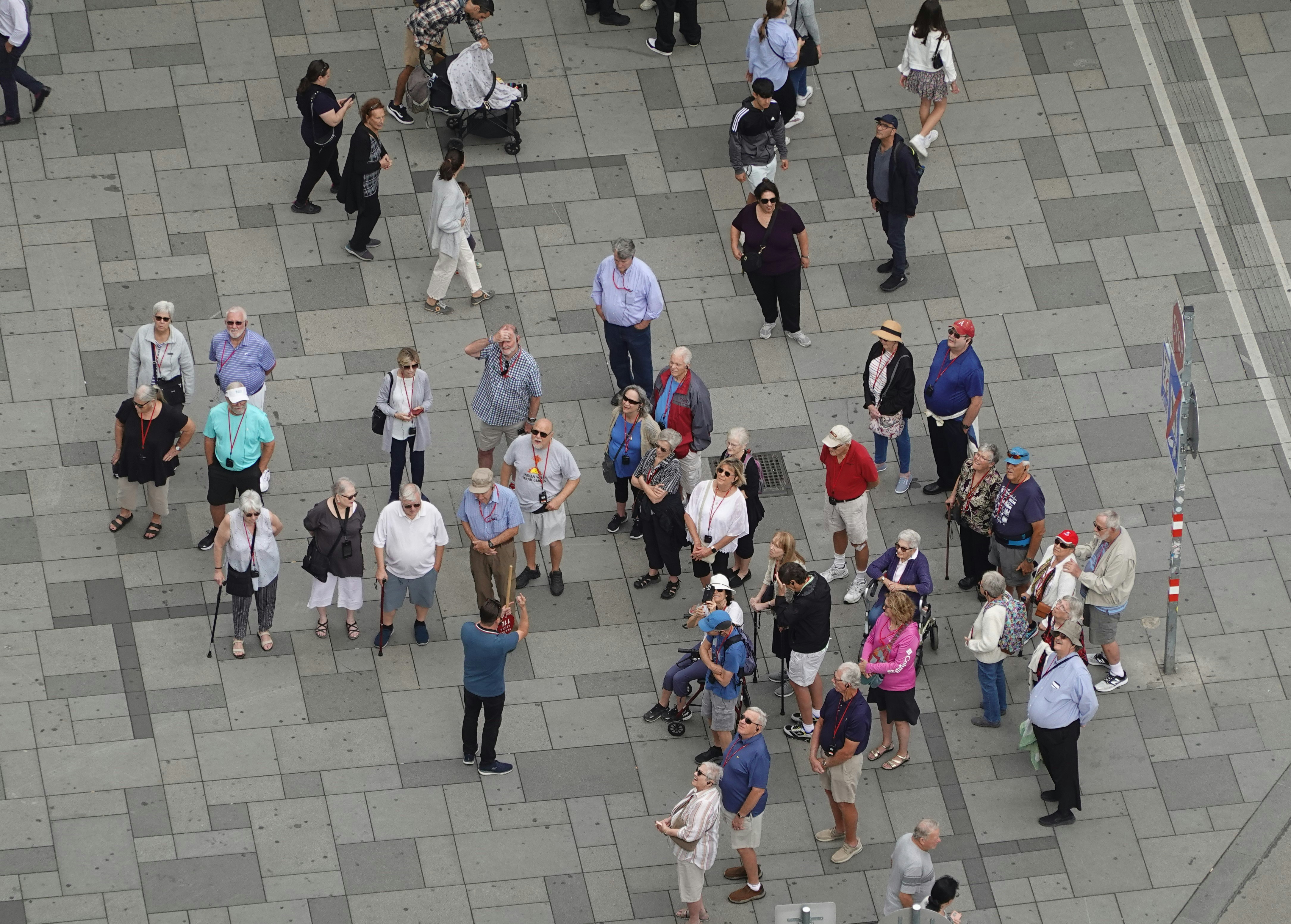 A group of people, primarily older adults, are gathered in what appears to be a public square or pedestrian area with tiled pavement. Some individuals are using walking aids or strollers, while others are casually standing or walking. The people are dressed in summer clothing, suggesting a warm day, and a few are wearing hats or sunglasses. The scene is viewed from above, showcasing a diverse group engaged in what might be a guided tour or a social gathering.