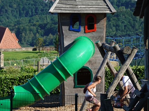 A wooden playhouse structure with a green enclosed slide stands in the foreground. Surrounding the playhouse, a jungle gym made of wooden logs and a blue rope bridge can be seen. Two adults and a child are interacting at the base of the structure. The background features a lush green landscape with forested hills and a building with a red roof.
