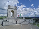 A large, historic stone monument with a central arch and decorative architectural elements. Two Italian flags are prominently displayed on flagpoles in the foreground. The monument is situated in a landscaped area with a mix of grass and stone paths, under a bright blue sky filled with fluffy white clouds. Rolling green hills are visible in the background.