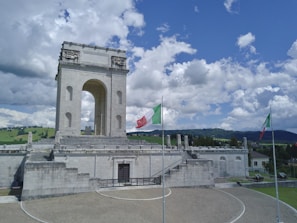 A large, historic stone monument with a central arch and decorative architectural elements. Two Italian flags are prominently displayed on flagpoles in the foreground. The monument is situated in a landscaped area with a mix of grass and stone paths, under a bright blue sky filled with fluffy white clouds. Rolling green hills are visible in the background.