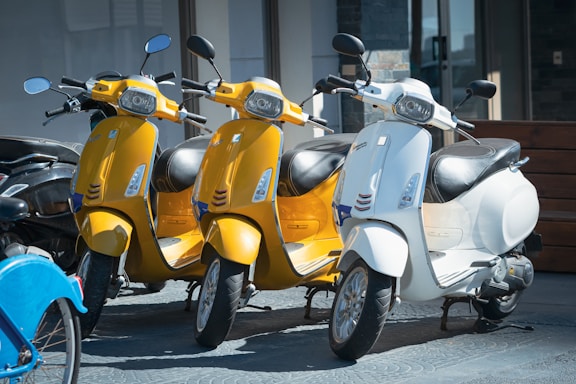 Three scooters, two in yellow and one in white, are parked closely together. The scooters have a classic design with round mirrors and prominent headlights. They are positioned on a textured pavement in front of a building with large windows. A blue bicycle's front wheel is partially visible in the foreground.