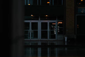 A dimly lit storefront with glass doors, reflecting the street in front. The neon sign displaying 'OPEN' is glowing in orange, providing the primary source of light. The scene is dark and moody, with wet reflections indicating recent rain. The building appears to be closed or not active despite the sign.