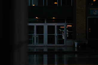 A dimly lit storefront with glass doors, reflecting the street in front. The neon sign displaying 'OPEN' is glowing in orange, providing the primary source of light. The scene is dark and moody, with wet reflections indicating recent rain. The building appears to be closed or not active despite the sign.