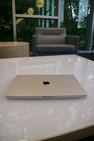 A silver laptop with an Apple logo is placed on a glossy white table. In the background, a grey armchair is positioned next to a round wooden side table. Large windows reveal greenery and hanging spherical lights outside.