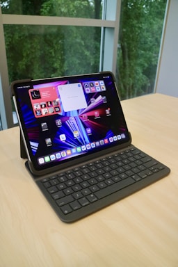 A tablet is positioned on a table with a detachable keyboard, displaying a colorful home screen with various app icons. The background shows a large window with green foliage visible outside, giving the setting a fresh and modern feel.