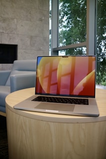 A sleek, modern laptop with a colorful screen is placed on a round wooden table. The background includes a light gray wall, a blue upholstered chair, and large windows with a view of green trees.