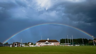 A vibrant softball field under a rainbow sky with players wearing colorful pride gear.