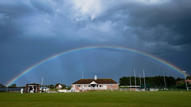 A vibrant softball field under a rainbow sky with players wearing colorful pride gear.