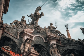 A detailed sculpture featuring several figures, including a unicorn, adorns a richly textured building facade. The craftsmanship includes a classical aesthetic with figures holding various objects, such as a lyre and a plant.