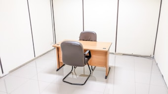 A minimalist office setting with a wooden desk and two brown leather chairs positioned facing each other. The room features plain white walls and a clean, tiled floor. There are visible sockets and wires on the floor against the wall.