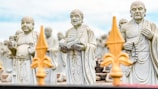 Three stone statues of Buddhist monks stand with distinct expressions and postures, surrounded by similar statues in the background. The central statue holds a book, and each monk is depicted with traditional robes and beads. The foreground features blurred decorative elements resembling fleur-de-lis designs, creating depth in the image.