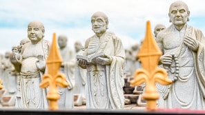 Three stone statues of Buddhist monks stand with distinct expressions and postures, surrounded by similar statues in the background. The central statue holds a book, and each monk is depicted with traditional robes and beads. The foreground features blurred decorative elements resembling fleur-de-lis designs, creating depth in the image.