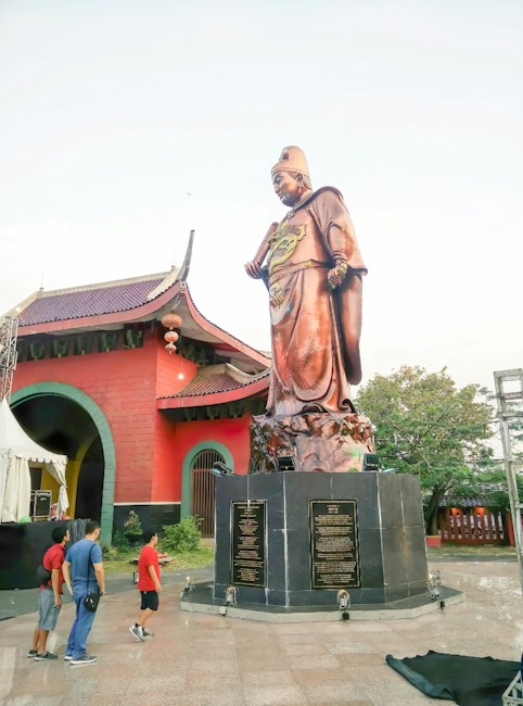 A large bronze statue of a historical figure stands prominently on a black pedestal with engraved plaques. The figure wears traditional clothing and a hat, with detailed patterns visible on the robe. In the background, there is an ornate building with a red facade and arched windows. A group of people is observed admiring the statue. The scene is set in a park-like area with trees visible in the surroundings.
