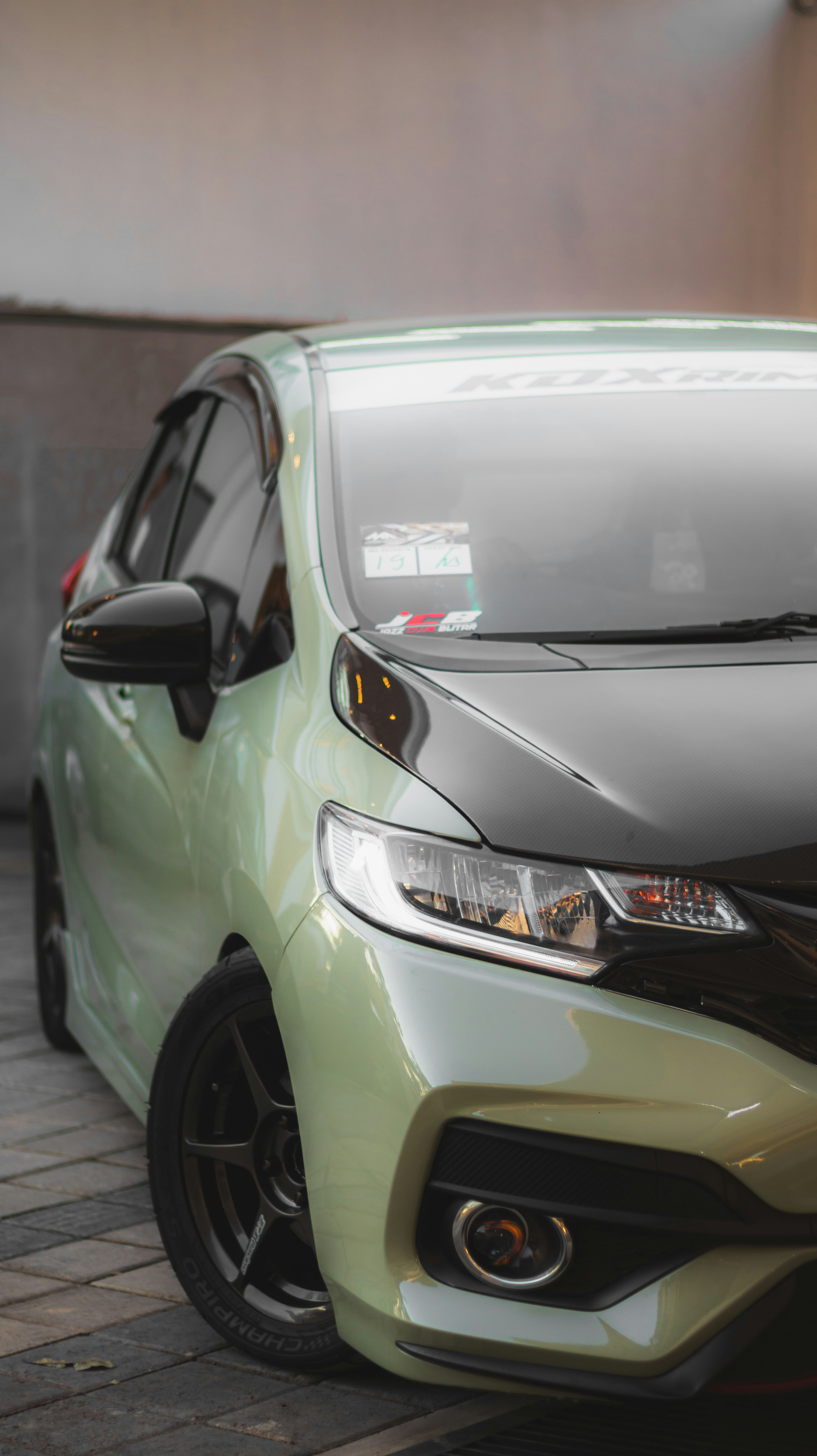 A sleek car with a modern design is parked on a tiled surface. The car features a shiny, light green exterior with a glossy black hood. It is equipped with stylish black wheels and a front grill with an integrated fog light. The windows are tinted, and there are decals on the windshield.