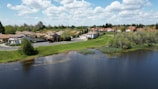 A suburban residential area with houses lining a tranquil street. A lush green park with trees and shrubs is adjacent to a calm lake. The sky is scattered with fluffy white clouds, creating a serene and peaceful atmosphere.
