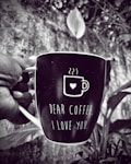 A hand is holding a coffee mug with a humorous and affectionate message on it. The black mug features a white illustration of a steaming coffee cup with a heart, and the words 'Dear Coffee, I Love You.' in white text. The background appears to be a natural setting with a blurred view of leaves and foliage, captured in black and white.
