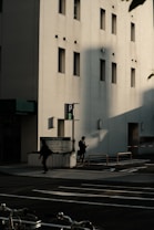 The image features an urban street scene with a modern building in the background. The building has a light-colored facade with several narrow windows. Shadows cast by the sun create dramatic shapes on the building's wall. In the foreground, two individuals are walking on the sidewalk, one of them appears to be looking at a phone. There is a parked bicycle partially visible at the bottom of the image.