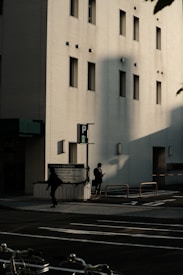The image features an urban street scene with a modern building in the background. The building has a light-colored facade with several narrow windows. Shadows cast by the sun create dramatic shapes on the building's wall. In the foreground, two individuals are walking on the sidewalk, one of them appears to be looking at a phone. There is a parked bicycle partially visible at the bottom of the image.