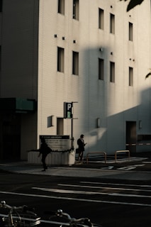 The image features an urban street scene with a modern building in the background. The building has a light-colored facade with several narrow windows. Shadows cast by the sun create dramatic shapes on the building's wall. In the foreground, two individuals are walking on the sidewalk, one of them appears to be looking at a phone. There is a parked bicycle partially visible at the bottom of the image.