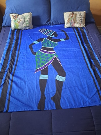 A bed is covered with a vibrant blue bedspread featuring an abstract design of a person in traditional attire. The figure is adorned with a patterned headdress and jewelry. Two decorative pillows sit at the head of the bed, each with illustrated designs and phrases on them.