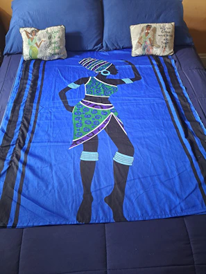 A bed is covered with a vibrant blue bedspread featuring an abstract design of a person in traditional attire. The figure is adorned with a patterned headdress and jewelry. Two decorative pillows sit at the head of the bed, each with illustrated designs and phrases on them.