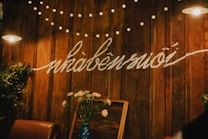 A cozy, warmly lit interior with wooden walls adorned with a string of small decorative lights and script writing. A vase with pink flowers sits on a table alongside a brown leather chair. The lighting creates a homely and inviting atmosphere.