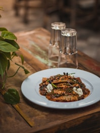 A rustic wooden table holds a white plate filled with a savory dish, possibly ravioli, garnished with herbs and dollops of cream. Nearby, two clear, inverted glasses stand on the table. To the left, a small green plant adds a touch of nature to the scene.