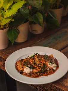 A rustic wooden table is adorned with potted green plants. At the center, a white plate holds a dish of ravioli, topped with rich tomato sauce and garnished with herbs and dollops of ricotta cheese.