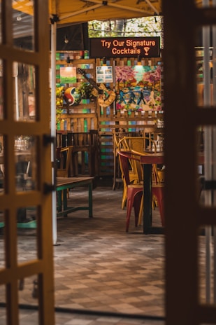 An inviting outdoor dining area with colorful decor, featuring various artwork and signs, including one advertising signature cocktails. The setting includes wooden tables and chairs, some painted in bright colors, and a partially open entryway providing a glimpse into the lively atmosphere.