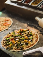 A freshly prepared pizza topped with a variety of colorful vegetables including peppers, broccoli, and olives. The pizza is placed on a counter next to another partially visible pizza. In the background, there are ingredient trays filled with toppings like olives and green peppers.