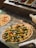 A freshly prepared pizza topped with a variety of colorful vegetables including peppers, broccoli, and olives. The pizza is placed on a counter next to another partially visible pizza. In the background, there are ingredient trays filled with toppings like olives and green peppers.