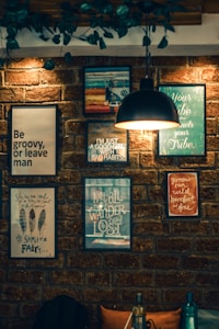 A brick wall decorated with a variety of framed motivational and inspirational quotes. The frames are of different sizes and colors, and a black pendant lamp hangs from above, casting a warm light. Lush green plants drape from the ceiling, adding to the cozy and artistic vibe.