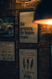 A dimly lit brick wall adorned with framed art and quotes, including a psychedelic van, a quote in large bold letters saying 'Be groovy, or leave man', and other smaller handwritten quotes relating to hippie culture. A warm light source is partially visible in the upper right corner.