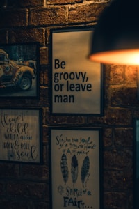 A dimly lit brick wall adorned with framed art and quotes, including a psychedelic van, a quote in large bold letters saying 'Be groovy, or leave man', and other smaller handwritten quotes relating to hippie culture. A warm light source is partially visible in the upper right corner.