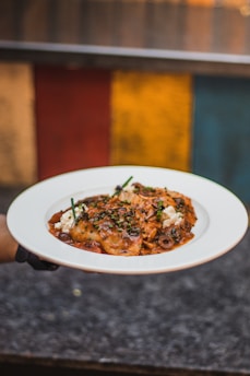 A white plate holds a dish with ravioli topped with sauce, garnished with herbs and possibly cheese, against a colorful background.