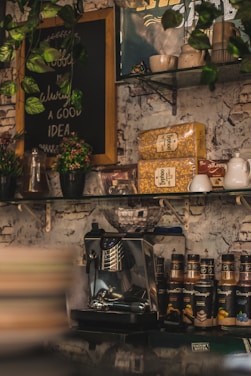 A cozy coffee shop corner featuring a coffee machine, various coffee syrups, and decoration items. There are green plants hanging and placed around, along with packaged coffee and a chalkboard sign with handwritten text. The background has a rustic, white-brick wall.