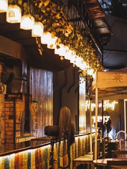 A cozy indoor setting with warm lighting and rustic d&eacute;cor, featuring a row of hanging pendant lights and foliage. A brick pizza oven is visible on one side, and a fan stands near the center. Neon signs glow in the background, contributing to a welcoming ambiance.