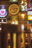 A cozy indoor setting with vibrant and eclectic decor. Various signs and advertisements for beer brands are visible on the walls, including neon signs and illuminated posters. There are hanging lights and plants adding warmth to the atmosphere.